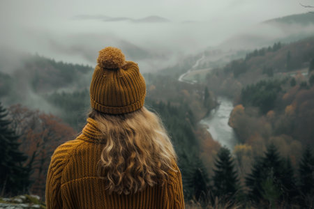 A woman stands overlooking a flowing river, her back to the viewer. She wears a brown knitted hat and a plaid jacket. The river flows through a dense forest with fog.の素材