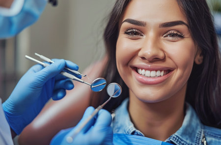 A woman smiles while a dentist uses a mirror to inspect her teeth. The dentist is wearing blue gloves and the woman is wearing a blue medical gown.の素材