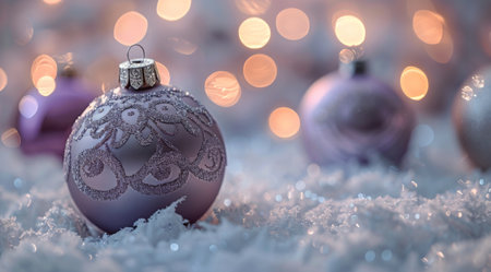 A close-up image of several purple Christmas ornaments resting on white faux snow. The ornaments have a shiny, sparkly finish and are decorated with intricate white patterns. The background is blurry and features a bokeh effect of warm white lights.の素材