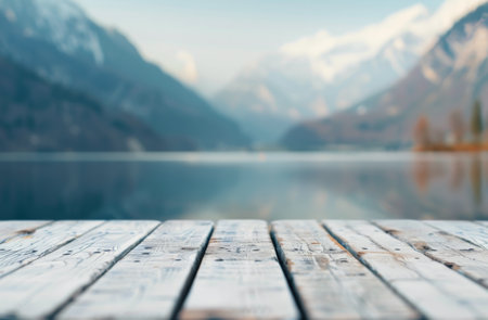A wooden dock extends out over a still lake, with a blurred background of mountains and trees. The surface of the water is calm and reflective.の素材