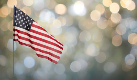A small American flag waves gently in the breeze, its red, white, and blue colors vibrant against the blurry backdrop of autumn foliage and sunlight.の素材