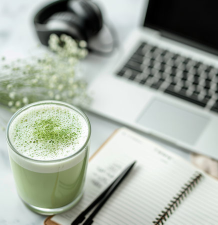 A close-up of a glass of matcha latte with a light foam layer, resting on an open notebook on a marble table. A laptop and headphones are visible in the background, suggesting a work or study environment.の素材