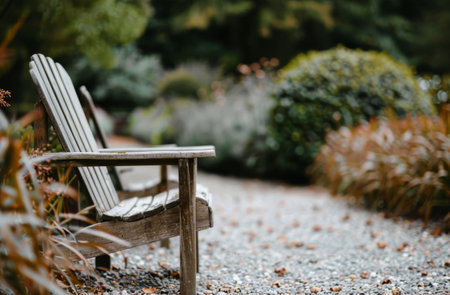 Two wooden chairs face each other on a gravel path, surrounded by lush greenery and soft, autumnal light. The chairs are empty, inviting viewers to imagine a peaceful moment spent in the garden.の素材