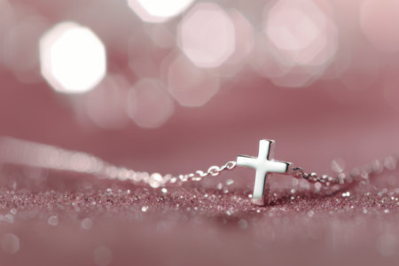 A silver cross necklace rests on a pink background with white flowers out of focus in the background.の素材