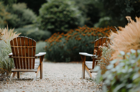 Two wooden chairs face each other on a gravel path, surrounded by lush greenery and soft, autumnal light. The chairs are empty, inviting viewers to imagine a peaceful moment spent in the garden.の素材
