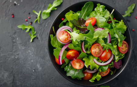 A white bowl filled with a vibrant green salad featuring spinach, cherry tomatoes, and red onion. The salad sits atop a dark gray surface.の素材