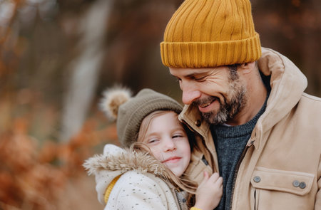 A young girl hugs her father from behind, both smiling brightly. They are in a forest setting, with autumn leaves visible in the background.の素材