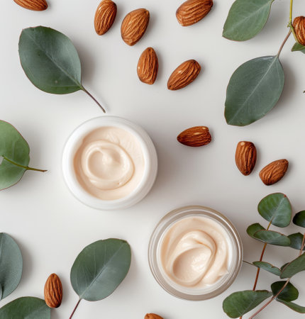 Two jars of creamy lotion are surrounded by almonds, eucalyptus leaves, and white flowers on a marble background. The jars are open, revealing the smooth, white cream inside.の素材