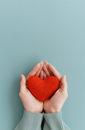 Two hands hold a red heart on a light blue background. The hands are positioned to gently cradle the heart, conveying a sense of care and affection.の素材