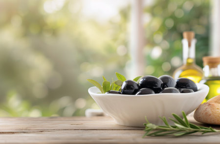 A white bowl filled with black olives sits on a wooden table with a sprig of rosemary in the foreground. The olives are surrounded by green leaves, and the background is a blurred image of a window with a view of green foliage.の素材