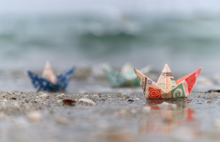 A paper boat made from a banknote floats on the wet sand of a beach. The boat is in focus, with the ocean and other paper boats in the background.の素材