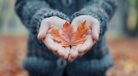 A individual stands in a forest during autumn, gently holding a bright orange maple leaf between their hands. The warm colors of the background complement the vivid leaf, capturing the essence of the season.の素材