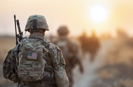 A group of soldiers march away, silhouetted against a vibrant sunset. Each carries a pack adorned with an American flag, highlighting their unity and purpose during a military operation.の素材