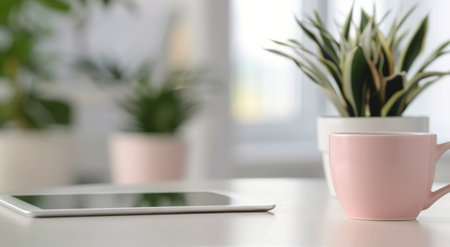 A soft pink mug sits next to a tablet on a clean desk, with vibrant green plants in the background, creating a tranquil workspace atmosphere filled with natural light.の素材