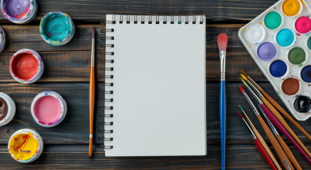 A sketch blankbook and a vibrant watercolor palette are placed on a rustic wooden table, inviting creativity and artistic expression. A paintbrush leans beside the open notebook, ready for use.の素材