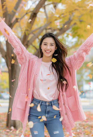 A young woman wearing a pink cardigan and light sweater joyfully throws autumn leaves into the air, surrounded by colorful fall foliage in a park, capturing a moment of happiness in the crisp afternoon.の素材