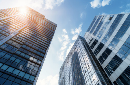 Sunlight beams brightly down from a clear blue sky, illuminating the towering glass skyscrapers of a modern city. The angles of the buildings create a dynamic visual experience against the sky.の素材