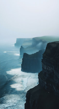The mysterious beauty of the Cliffs of Moher is captured as waves crash against the rocky shoreline. Mist cloaks the landscape, enhancing the dramatic view of the cliffs.の素材