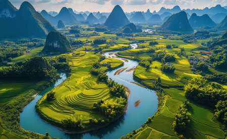 Lush verdant rice terraces cascade down hillsides alongside a serpentine river, set against a backdrop of sweeping mountain ranges in Guangxi province during daylight.の素材