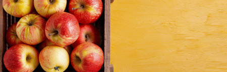 A wooden crate filled with vibrant red and yellow apples sits against a sunny yellow backdrop, showing the fresh harvest. The apples display a natural sheen, highlighting their freshness and quality.の素材