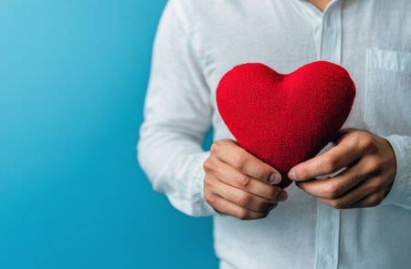 A person with a white shirt holds a red heart-shaped cushion close to their chest against a vibrant blue background, conveying warmth and affection in a cozy indoor setting.の素材