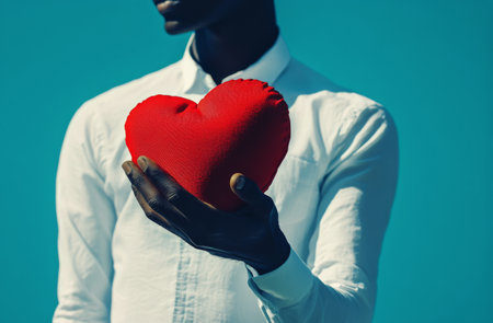 A person with a white shirt holds a red heart-shaped cushion close to their chest against a vibrant blue background, conveying warmth and affection in a cozy indoor setting.の素材