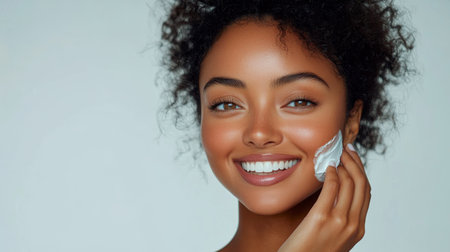 A cheerful woman uses a creamy skincare product on her face, showing healthy skin. Her curly hair frames her warm expression, highlighting her enjoyment in self-care.の素材
