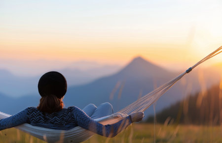 A woman reclines in a hammock, facing away from the camera, and looks out towards a picturesque mountain range as the sun sets.の素材