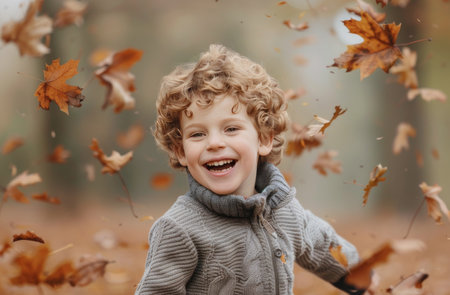 A young boy with curly blonde hair laughs as he stands in a field of falling autumn leaves. The leaves are a warm brown and orange color, and the boy is wearing a gray sweater.の素材