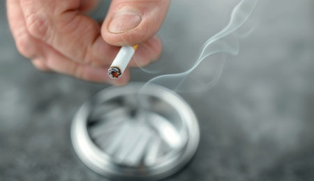 A hand holds a lit cigarette over an ashtray. Smoke rises from the cigarette, forming wispy patterns in the air.の素材