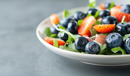 A close-up view of a salad with blueberries, leafy greens, and prosciutto in a white bowl.の素材