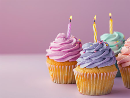 Three colorful cupcakes with candles, decorated with frosting and sprinkles, on a pink surface.の素材