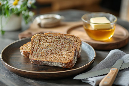 A rustic wooden board showcases freshly baked whole grain bread with oat flakes. Slices are laid out beside a jar of honey and butter, creating a warm, inviting breakfast setting.の素材