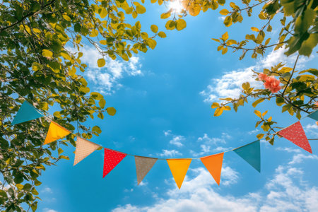 Brightly colored bunting flags are strung between leafy branches in a park. The sun shines warmly, casting a cheerful atmosphere on a beautiful day with a few fluffy clouds.の素材