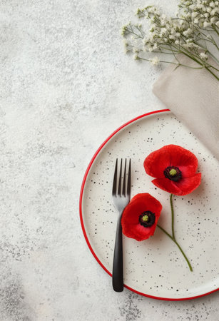 Two vibrant red poppy flowers rest on a simple white plate with a red rim, accompanied by a fork and napkin. The setting features a textured gray background that enhances the colors.の素材