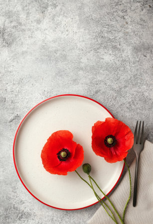 Two vibrant red poppy flowers rest on a simple white plate with a red rim, accompanied by a fork and napkin. The setting features a textured gray background that enhances the colors.の素材