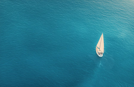 A white sailboat glides smoothly across deep blue waters, creating gentle ripples. The sunlight reflects on the surface, enhancing the serene atmosphere of a perfect sailing day.の素材