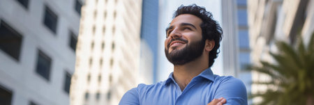 A young man with a beard stands confidently with his arms crossed, smiling against a backdrop of tall modern buildings under a clear blue sky.の素材