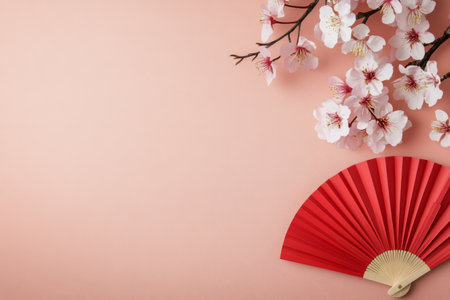 Delicate cherry blossom branches with pink flowers lay beside a stylish red fan on a light pastel background. This composition evokes a sense of tranquility and beauty, perfect for seasonal themes.の素材