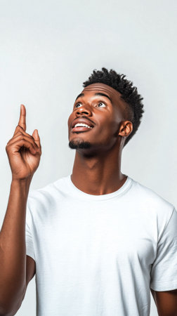 A young man is standing against a plain background while pointing upwards with a delighted expression. He is wearing a plain white t shirt, highlighting his cheerful demeanor.の素材