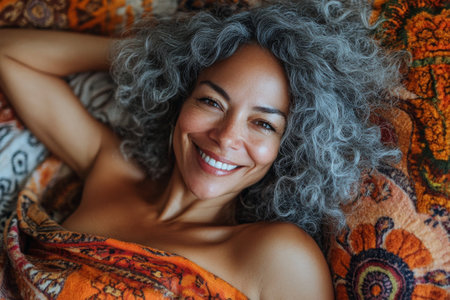 A woman with long, wavy hair relaxes on a vibrant blanket. She smiles warmly, surrounded by rich colors and a peaceful atmosphere during the evening light.の素材