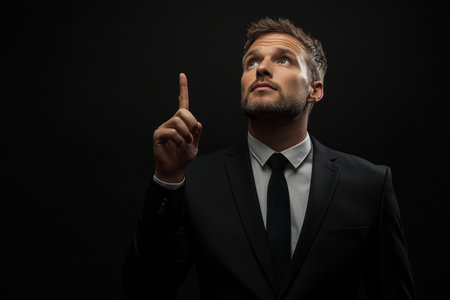 A businessman dressed in a black suit and tie looks up, raising one finger as if making a point. The dark background creates a serious atmosphere, highlighting his expression.の素材