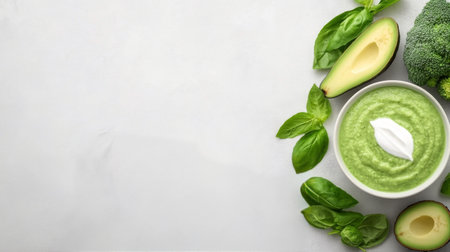 A bowl of creamy green soup made with broccoli and avocado is garnished with a dollop of cream. Fresh basil leaves and half an avocado surround the bowl on a light gray surface.の素材