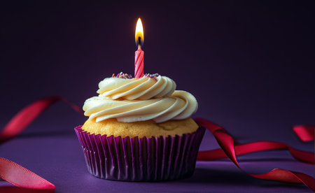 A beautifully decorated cupcake sits against a rich purple background. Topped with creamy frosting and a lit pink candle, it is surrounded by vibrant red ribbons, perfect for celebrations.の素材