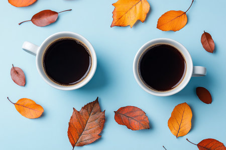 Two white coffee cups filled with dark coffee are placed on a light blue surface, surrounded by vibrant autumn leaves in various hues of red and orange, evoking a cozy atmosphere.の素材