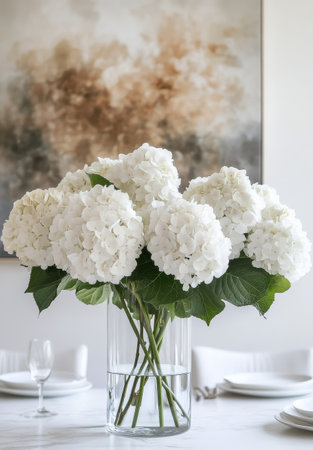 A vibrant display of white hydrangeas fills a clear glass vase, enhancing a sleek dining table. The soft colors of the surrounding artwork complement the floral arrangement perfectly.の素材