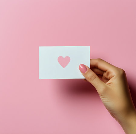 A hand with a manicured pink nail holds a white card featuring a heart shape cutout against a soft pink backdrop. This showcases a minimalist design concept.の素材