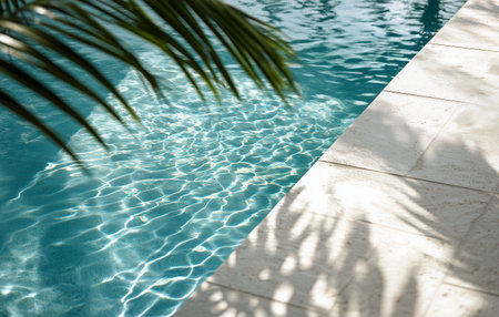 The crystal-clear water of a swimming pool reflects sunlight, while palm leaves create gentle shadows along the white stone edge in a serene outdoor environment.の素材