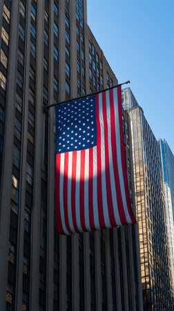A large American flag hangs from a high-rise building in a city, fluttering gently in the wind. The clear blue sky enhances the flags vibrant colors, surrounded by modern architecture.の素材