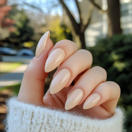 A hand displaying long almond-shaped nails painted in a nude color. The background features lush greenery and soft sunlight, emphasizing the nail designs elegance.の素材
